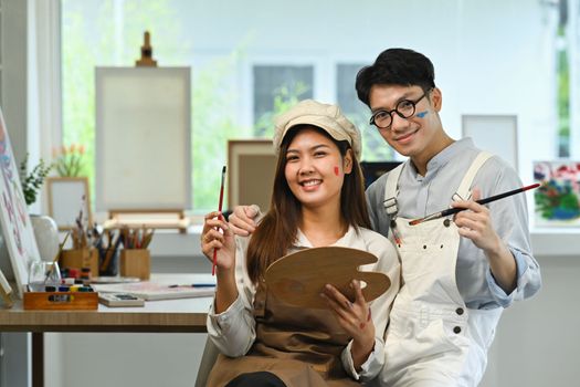 Young couple with paint brush and palette in hands sitting in bright art studio. Leisure activity, creative hobby and art concept. 