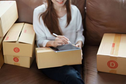 Starting small business entrepreneur of independent young Asian woman online seller is using tablet computer and taking orders to pack products for delivery to customers. SME delivery concept.