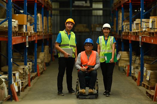 Image of middle age male manager in wheelchair and young workers standing in the retail warehouse full of shelves with merchandise.