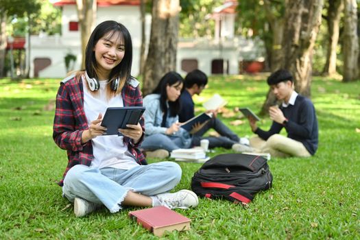 Cheerful asian female college or university student using digital tablet on green grass in the campus.