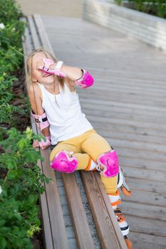 A little Caucasian girl in roller skates sits on a bench