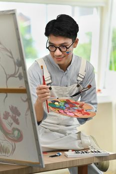 Happy asian male artist holding paintbrushes and palette painting on canvas at art studio. Education, hobby, art concept.