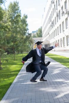 Old happy man in graduation gown jumping outdoors and holding diploma. Vertical