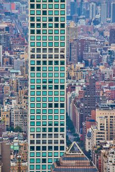 Image of Tall narrow skyscraper of turquoise squares with background of small buildings from above vertical