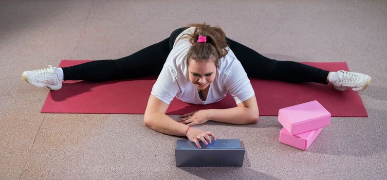 Young caucasian fat woman doing bends on a sports mat and watching a training video on a laptop. A chubby girl stretches the split remotely using video communication.