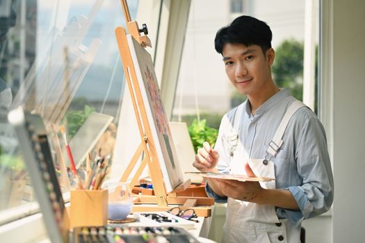 Happy asian male artist holding paintbrushes and palette painting on canvas at art studio. Education, hobby, art concept.