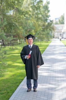 An old man in a graduation gown walks outdoors and holds a diploma