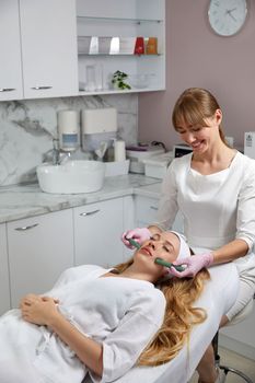 Young woman receiving facial massage with stone sticks in beauty salon, concept of face massage in spa
