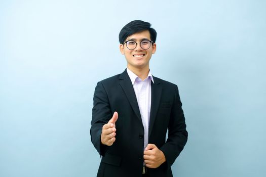 Portriat of asian young smart happy businessman dressed in suit standing straight, smiling, and pointing finger at camera with isolated light blue background. Business concept.