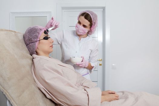 Carbon peeling in beauty salon. Cosmetologist applying black mask on the face of beautiful woman for carbon peeling