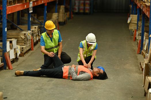 Two young storehouse colleagues helping middle aged worker lying on the floor in warehouse. Safety first concept.