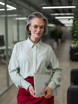 Mature caucasian woman stands with a laptop among the office