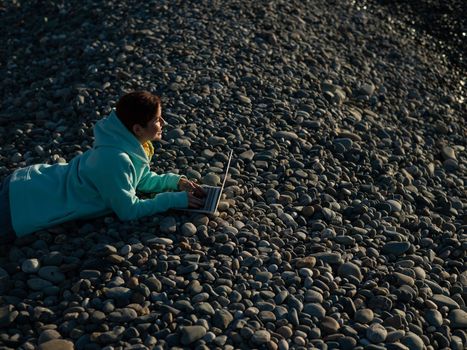 Caucasian woman lying on her stomach on a pebble beach and typing on a laptop. Remote work freelancer