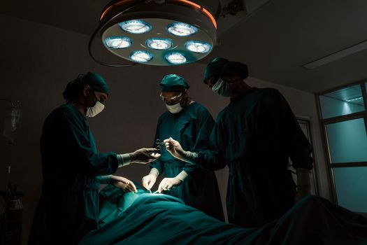 Surgical team performing surgery to patient in sterile operating room. In a surgery room lit by a lamp, a professional and confident surgical team provides medical care to an unconscious patient.