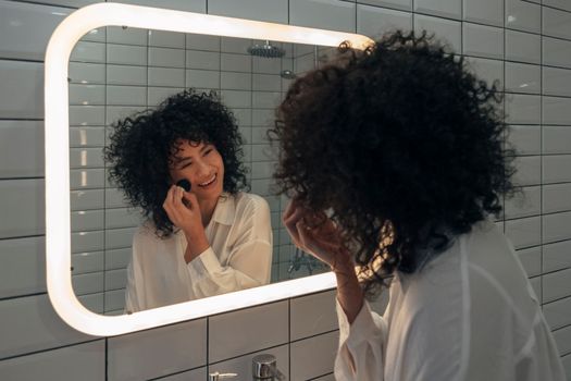 Young hispanic afro woman applying make-up with brush looking herself in the mirror laughing. Beauty concept. Getting ready concept.