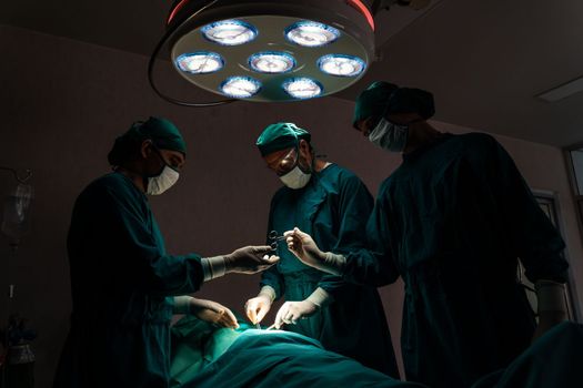 Surgical team performing surgery to patient in sterile operating room. In a surgery room lit by a lamp, a professional and confident surgical team provides medical care to an unconscious patient.