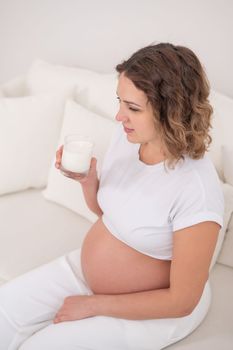 A pregnant woman sits on a white sofa and drinks milk