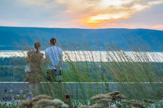 A couple standing on the edge of the observation deck, watching a beautiful sunset from a height. Summer abstract nature background with grass in a meadow and a sunset sky. Natural landscape.