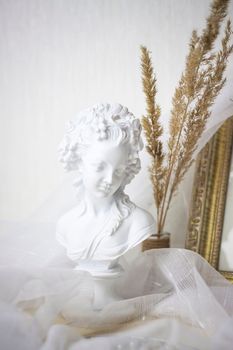 Plaster bust of a girl. Statuette on the background of light fabric and fluffy branches.  Selective focus
