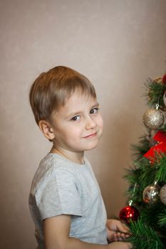 A photo of a beautiful boy in a gray T-shirt at the Christmas tree, looking into the camera. Portrait in a bright room. Natural, not staged photography. Children