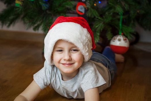 A photo of a beautiful boy in a gray T-shirt and a Santa Claus hat at the Christmas tree, looking into the camera. Portrait in a bright room. Natural, not staged photography. Children