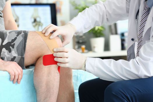 Physiotherapist applying kinesiotape to patient knee in clinic closeup. Injury and sprain of knee joint concept