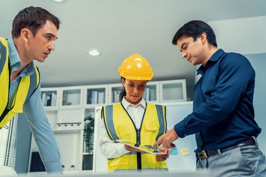 A team of competent engineers wearing safety equipment is working on blueprints with a tablet while also discussing with the investor in the office.