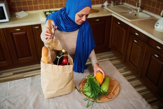 View from above of a Middle-Eastern Muslim woman in blue hijab and casual clothes, sorting fresh greens, organic vegetables while unpacking grocery shopping bag in the kitchen at home