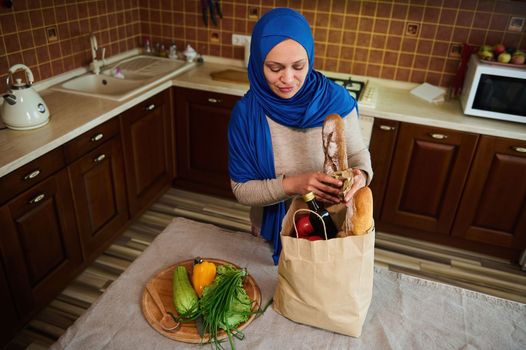 Overhead view of a Muslim Middle-Eastern pretty woman in blue hijab, removing groceries from a shopping bag, in the kitchen at home. Healthy grocery purchasing. Food delivery.