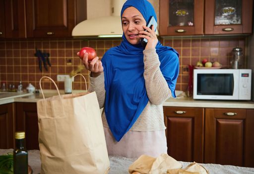 Pleasant housewife, Arab Muslim woman in blue hijab, returning home from grocery store, talking on mobile phone, taking fresh vegetables out of bags, unpacking groceries in the kitchen. Food delivery