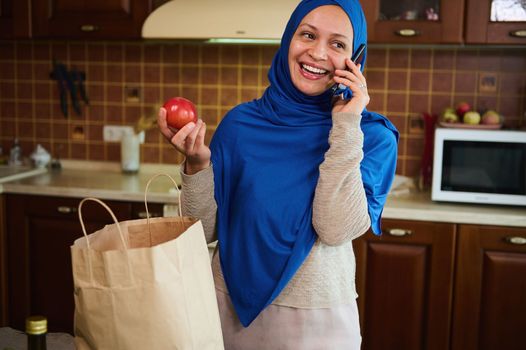 Charming Muslim woman from the Middle East speaks emotionally on her mobile phone, taking out purchases from grocery bags. Pretty Arab housewife with head in hijab smiles while unpacking shopping bags