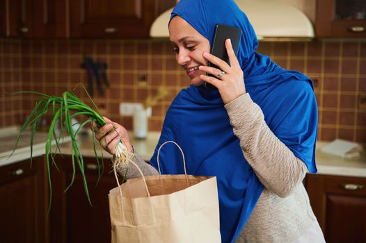 Young adult Arabic Muslim woman wearing a blue hijab, talking on mobile phone, unpacking eco shopping bag with fresh food, starting to lay out vegetables on the table in the home cozy wooden kitchen