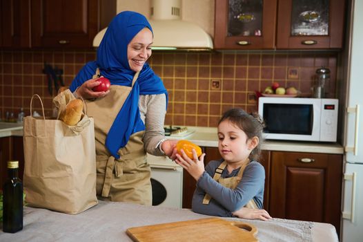Middle-Eastern Muslim cute woman in hijab, puts a bag of groceries on the table. Little girl helps her loving mom unpacking shopping bags with healthy food in the kitchen. Eco-friendly paper packaging