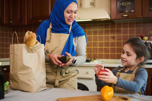 Caucasian little girl helps her mother unpack the delivered vegetables, takes out bell pepper, fresh clean paprika from paper bag. Muslim woman and her daughter arrive home after grocery shopping