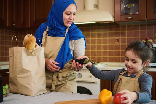 Cheerful happy Middle Eastern Muslim woman housewife in blue hijab, a loving mother and her lovely daughter unpacking grocery bag after shopping. Customers of online supermarkets. Food delivery