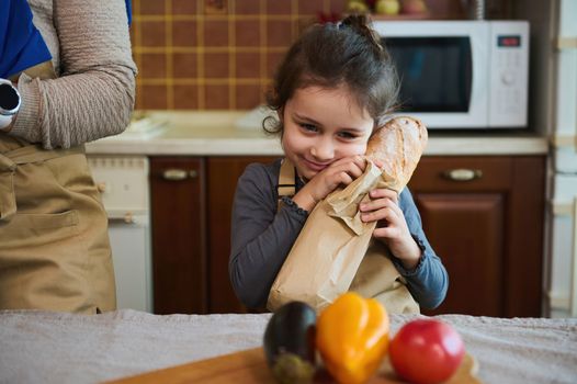 Adorable child, cute baby girl in a beige chef's apron, holds a hot baguette of freshly baked whole grain sourdough bread, while helping her mom unpacking shoppng bag in the kitchen with rustic design