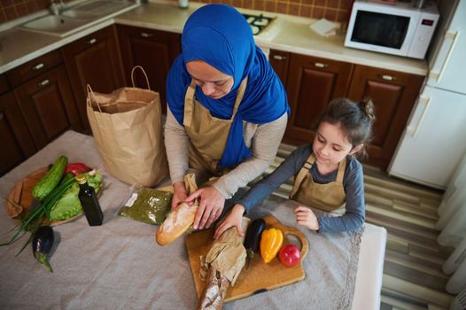 Top view. Happy Middle Eastern family relationship at home. Arab Muslim woman, loving mom with little daughter unpacking food, vegetable and fruit from supermarket bag together for preparing meal