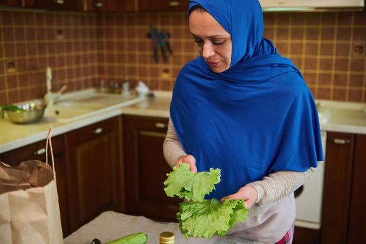 Middle-Eastern Muslim woman, a pretty housewife with head covered in blue hijab, holds fresh salad leaves in her hands while unpacking grocery bag in the home kitchen, after shopping. Food delivery.