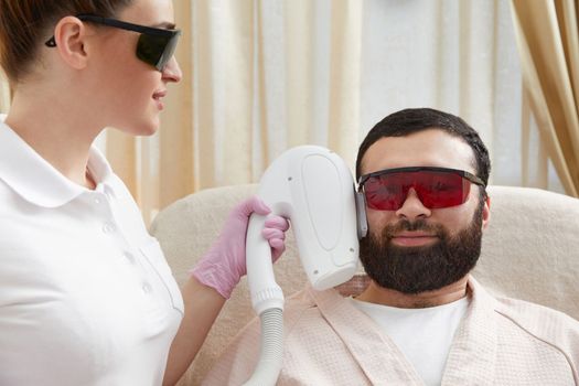 Bearded man getting laser facial treatment by professional cosmetologist in a beauty clinic. Healthy man lifestyle concept