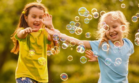 Two cheerful cute girls are playing outdoors with huge soap bubbles.