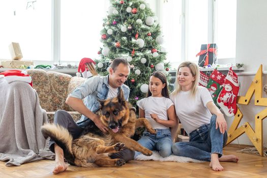 Friendly family is playing with dog near Christmas tree. They are sitting and laughing.