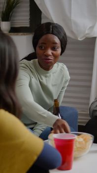 Afro american woman talking with friends enjoying time spend together during nightlife party. Group of mixed race people sitting on sofa, drinking beer late at night in living room