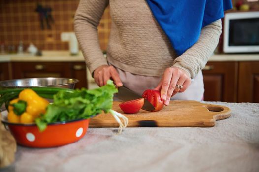 Cropped view: housewife in blue hijab, using kitchen knife, slicing tomatoes on a wooden cutting board, preparing raw vegan salad from fresh organic vegetables in the kitchen at home. Healthy eating