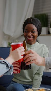 Afro american woman talking with her friends holding beer bottle while sitting on sofa late at night in living room. Group of mixed race people hanging out enjoying time spending together