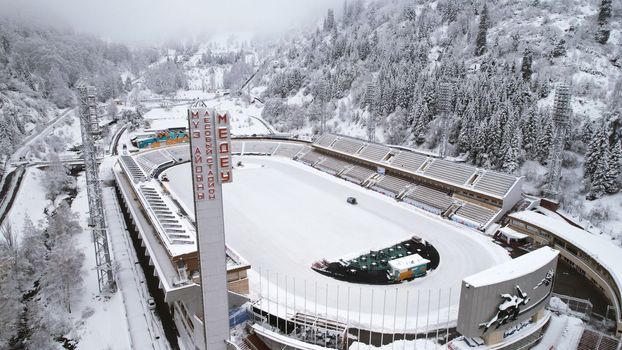 Winter alpine skating rink Medeo in the mountains. Drone view of the snowy forest and mountains. White clouds and fog in the gorge. Snow removal is underway. A large stadium. Almaty, Kazakhstan
