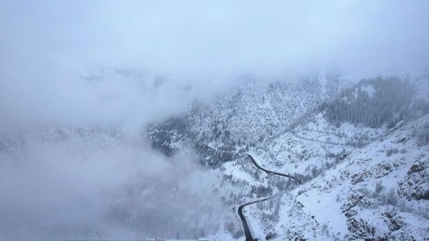 Snowy mountains with coniferous trees in the clouds. Medeo Dam. Everything is in fog and snow. Christmas and New Year have come. Aerial view from the drone on road, dam and trees. Almaty, Kazakhstan