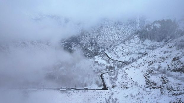 Snowy mountains with coniferous trees in the clouds. Medeo Dam. Everything is in fog and snow. Christmas and New Year have come. Aerial view from the drone on road, dam and trees. Almaty, Kazakhstan