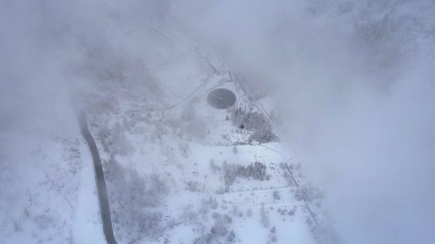 Snowy mountains with coniferous trees in the clouds. Medeo Dam. Everything is in fog and snow. Christmas and New Year have come. Aerial view from the drone on road, dam and trees. Almaty, Kazakhstan