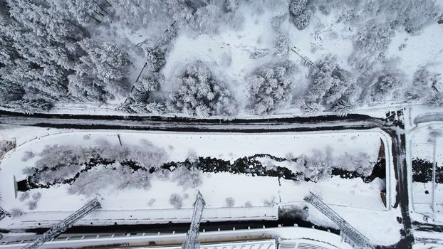 Snowy fairy-tale road in a mountain forest. Christmas or New Year has come. Coniferous trees in the snow. The river runs, next to the tall building of the Medeo skating rink. The view from the drone