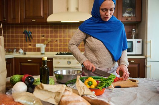 Charming Middle-Eastern Muslim woman, pretty housewife in blue hijab, standing by a kitchen table, chopping tomatoes on a cutting board, preparing delicious healthy salad from organic fresh vegetables